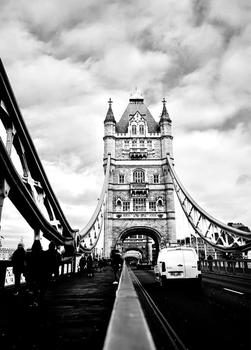 Gotische Türme der Tower Bridge vor bewölktem Himmel, ein Lieferwagen fährt über die Straße darunter.
