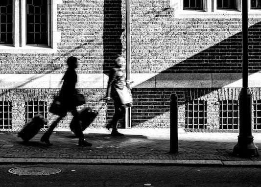 Black-and-white street photo of two blurred pedestrians pulling suitcases