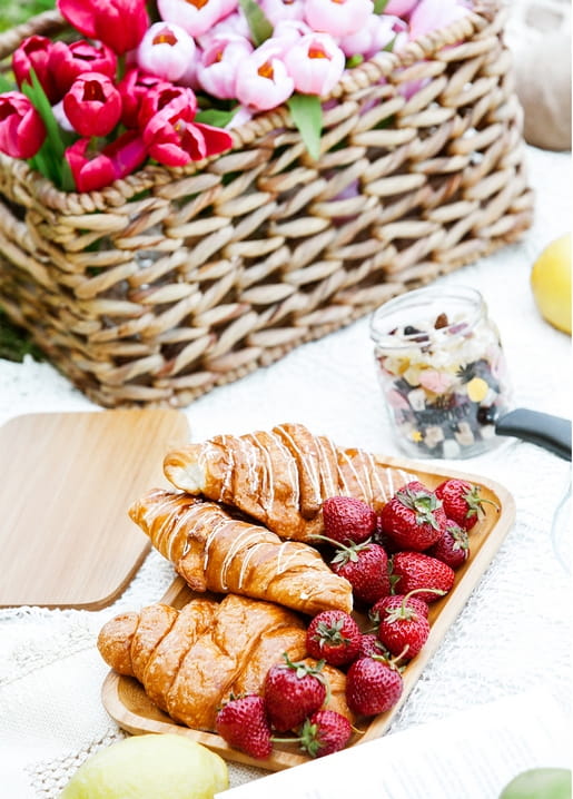 Wicker basket of red and pink tulips beside croissants and strawberries on a wooden board