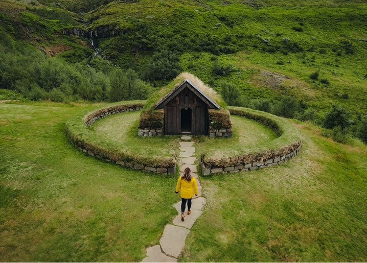 Hiker in a yellow raincoat walking toward a turf-roofed cottage ringed by a stone wall