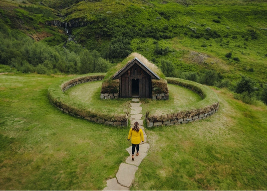 Hiker in a yellow raincoat walking toward a turf-roofed cottage ringed by a stone wall