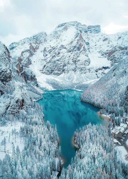 Aerial view of a turquoise alpine lake surrounded by snowy pine forest and mountains