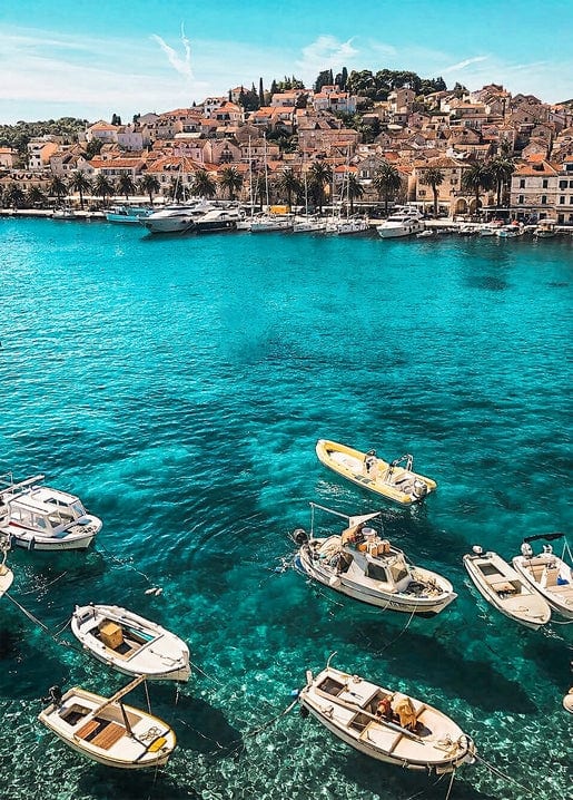 Small wooden fishing boats anchored in turquoise water below a sun-lit Mediterranean hillside town.