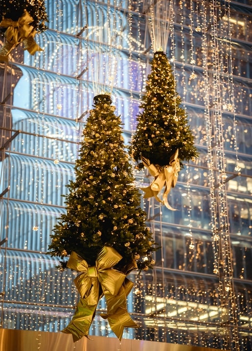 Two Christmas trees hanging upside-down with gold bows in front of a glowing glass building facade
