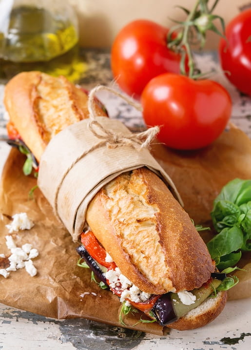 Baguette-Sandwich in Packpapier neben Tomaten am Zweig, Basilikum und Olivenöl