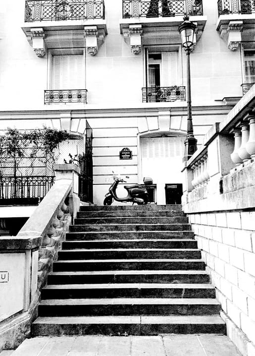 Ein Roller steht am Fuß einer Steintreppe unter schmiedeeisernen Balkonen in einer Pariser Straße.