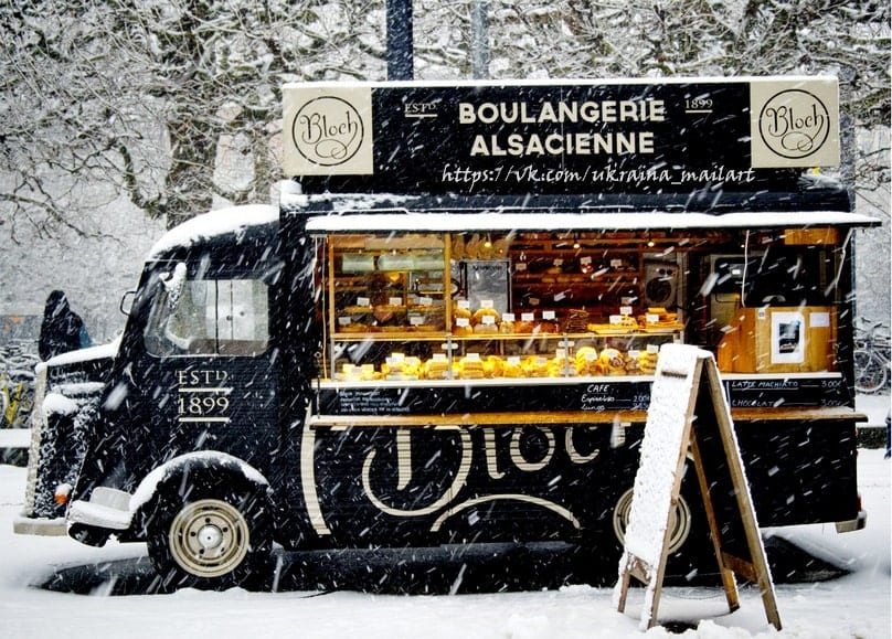 Schwarzer Vintage-Bäckerwagen mit hell erleuchtetem Schaufenster auf verschneiter Straße bei Schneefall