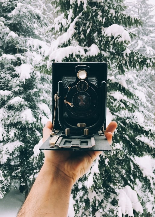 Hand holding an antique folding bellows camera against snow-covered pine trees