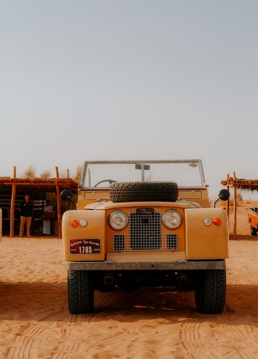 Vintage yellow Land Rover parked on a sandy beach with thatched huts behind