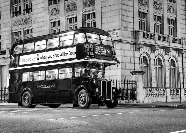 A vintage London double-decker bus glowing under night lights outside a grand building, black-and-white postcard.