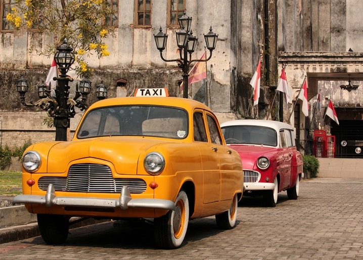 Gelbes Vintage-Taxi und roter Oldtimer vor einem verwitterten Gebäude mit Fahnen