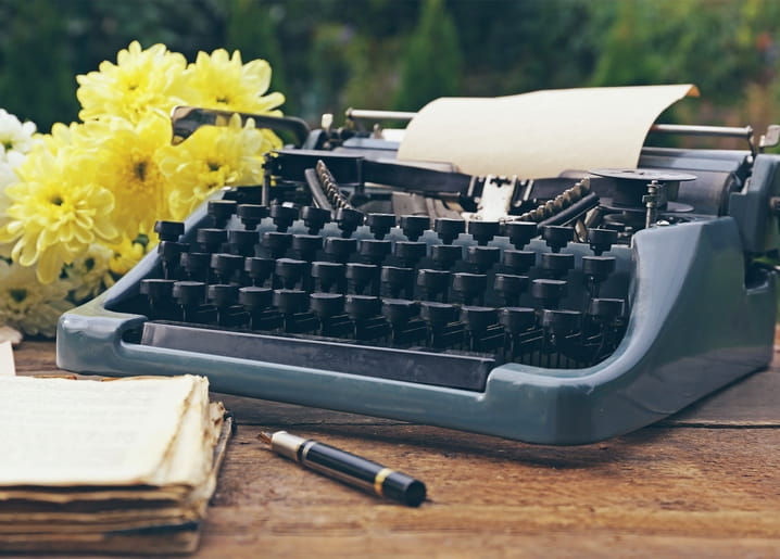 Alte Schreibmaschine mit gelben Chrysanthemen, einem Buch und einem Füllfederhalter auf einem Holztisch