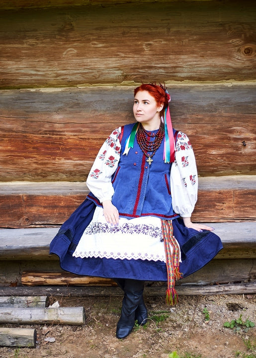 Red-haired woman in embroidered vyshyvanka and blue vest leaning against a log cabin wall
