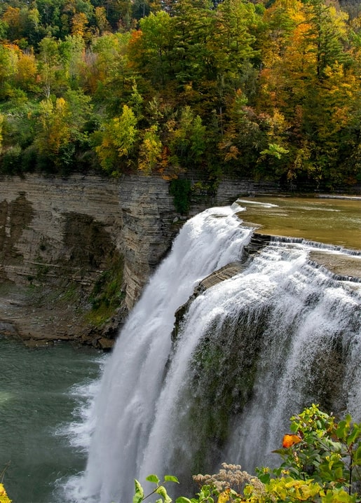 Breiter Wasserfall über gestuften Felsen, umgeben von herbstlich gefärbtem Wald