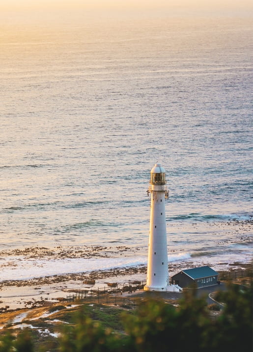 White lighthouse glowing in golden sunset light above a rocky coastline