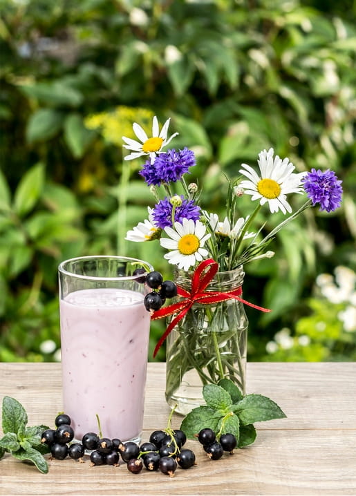 Glas mit Gänseblümchen und Kornblumen neben einem rosa Johannisbeer-Smoothie und verstreuten Beeren auf einem Holztisch