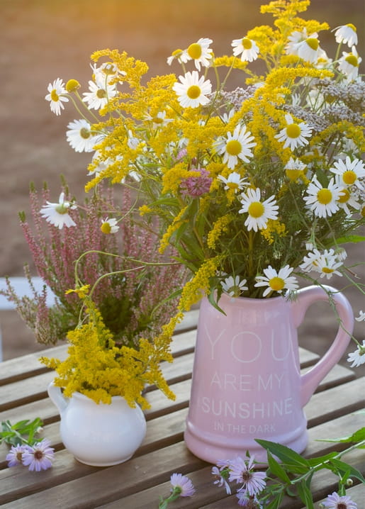 Daisies and goldenrod in a pink pitcher beside a small jug of pink heather on a wooden table