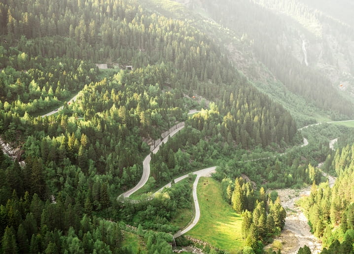 Luftaufnahme einer Serpentinenstraße durch ein neblig-grünes, bewaldetes Bergtal