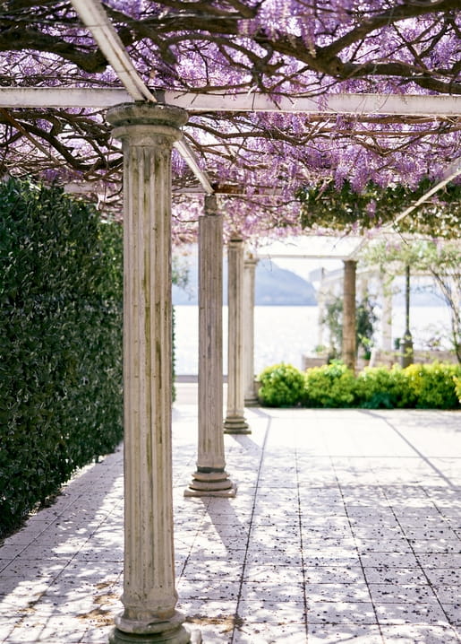 Stone pergola covered in purple wisteria with a garden path overlooking a lake