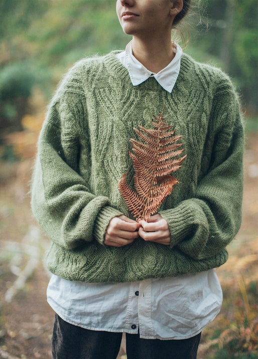 Woman in a green knit sweater holding a coppery fern leaf in a misty forest