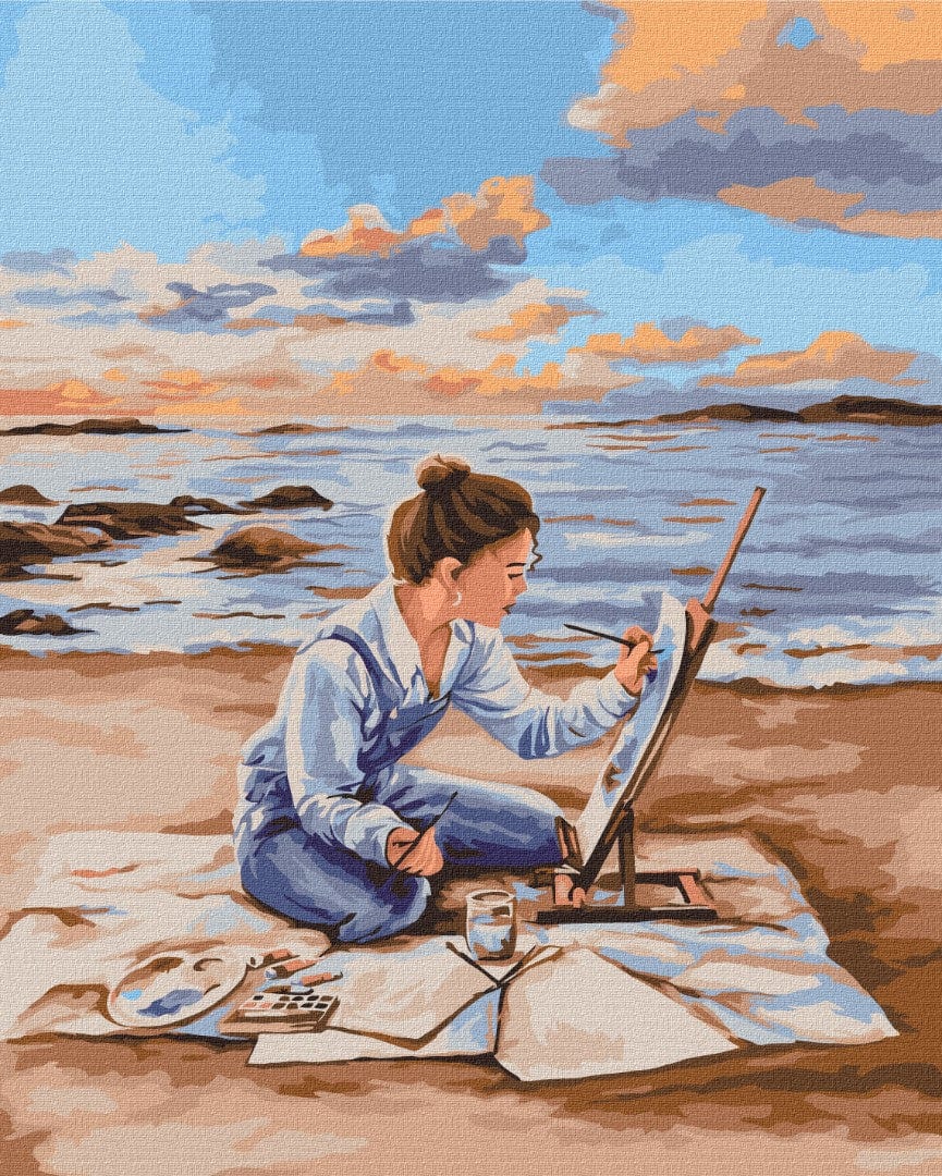 Frau im Jeansoverall malt einen Sonnenuntergang am Meer, sitzend auf einer Stranddecke
