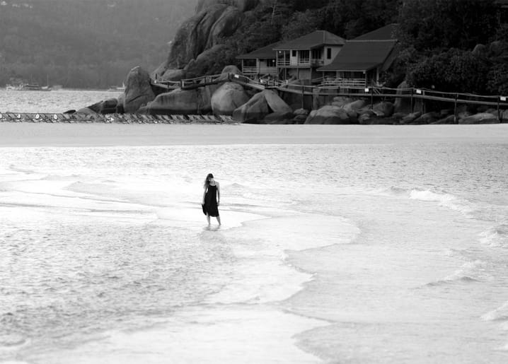 Woman in a dark dress walking along the shoreline at dusk, wooden lodge on rocks behind her