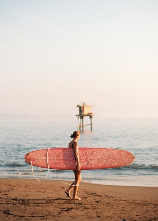 Woman walking a beach at sunset carrying a pink surfboard, lifeguard tower behind
