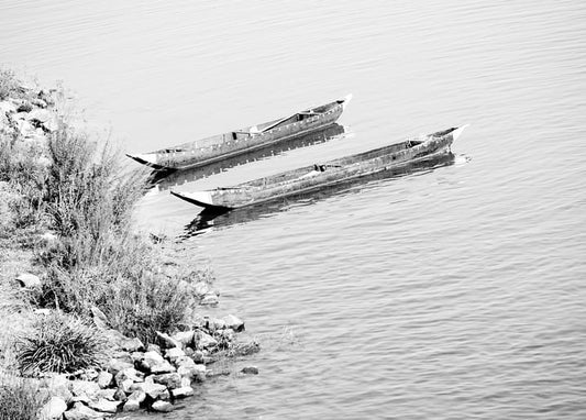 Two weathered wooden boats beached side by side on a calm, reed-lined shore, black-and-white