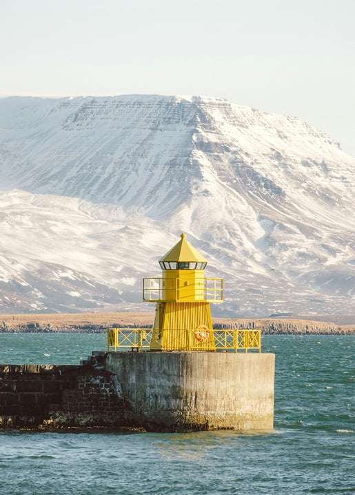 Yellow lighthouse on a stone pier with a snow-capped mountain behind