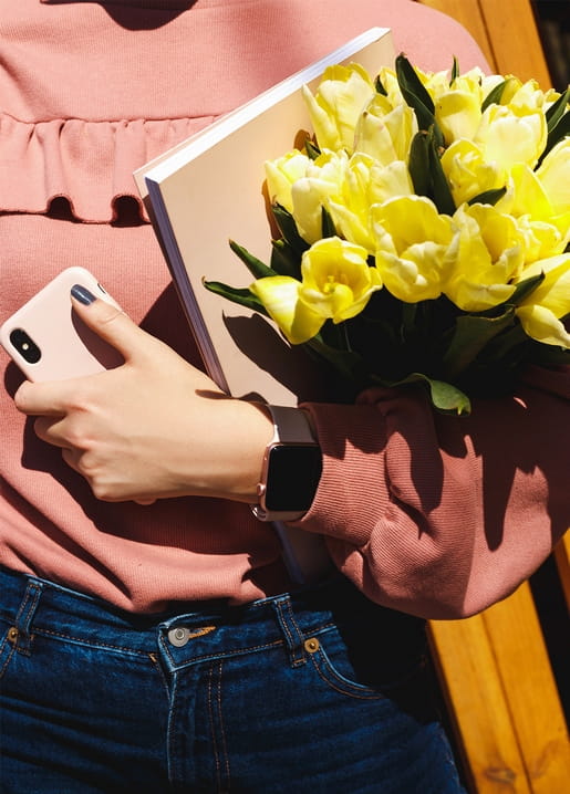 Woman in a pink sweater holding a book and a bouquet of yellow tulips, phone in hand