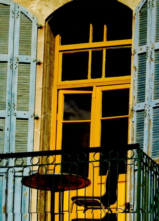 Glowing yellow window and door framed by weathered blue-grey shutters and an iron balcony railing.