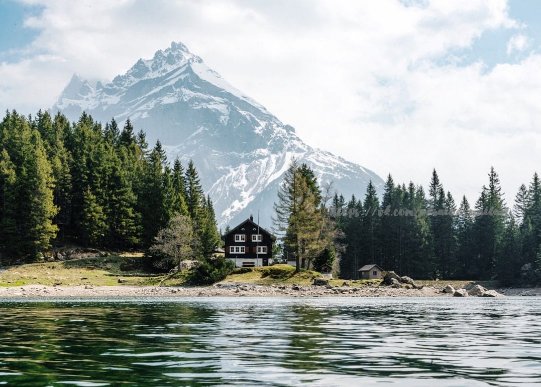 Schneebedeckter Berggipfel über einem Alpensee mit dunkler Holzhütte und Kiefernwald am Ufer