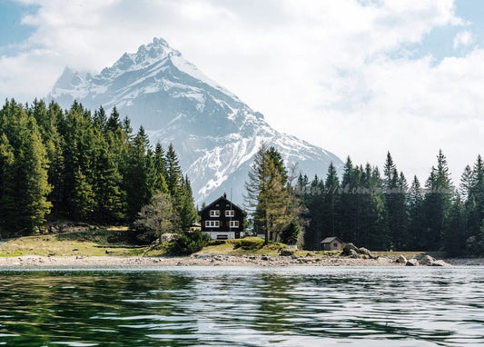 Schneebedeckter Berggipfel über einem Alpensee mit dunkler Holzhütte und Kiefernwald am Ufer