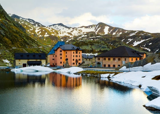 Ockerfarbene Alpengebäude mit dunklen Dächern spiegeln sich in einem See vor schneebedeckten Gipfeln.