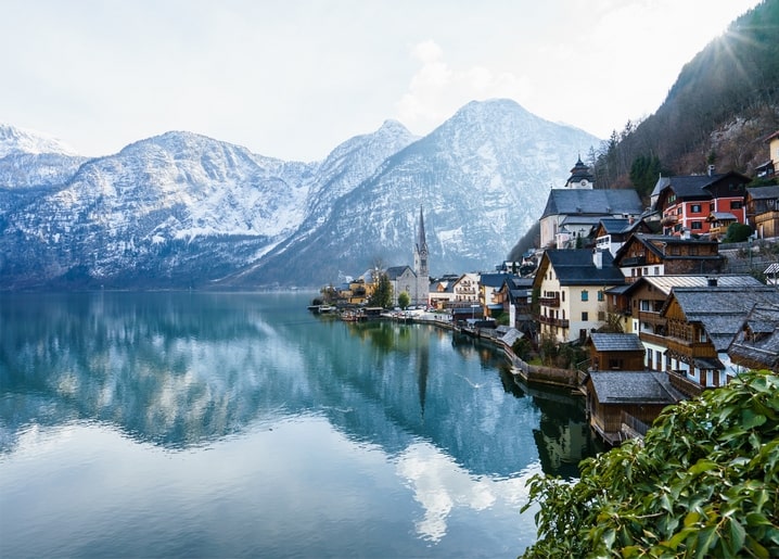 Alpines Dorf mit Kirchturm, gespiegelt im Wasser unter schneebedeckten Gipfeln