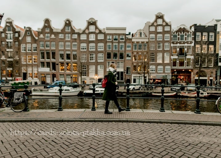 Frau im grünen Mantel spaziert an einer Amsterdamer Gracht mit Giebelhäusern