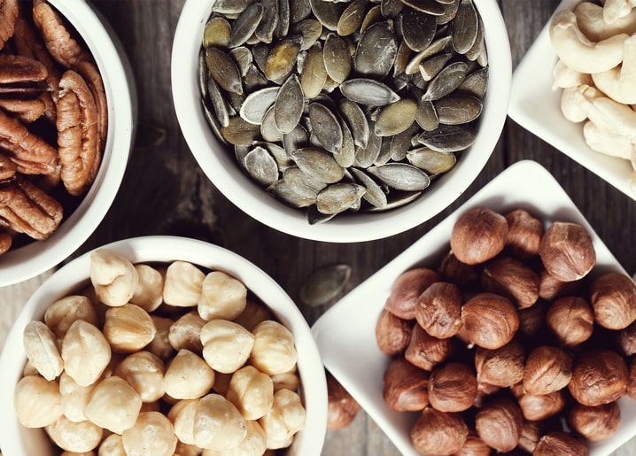Overhead flat lay of pecans, pumpkin seeds, cashews and hazelnuts in white bowls