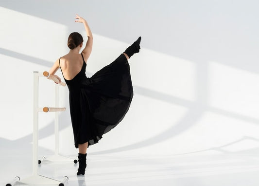 Ballerina im schwarzen Kleid von hinten, das Bein an der Ballettstange ausgestreckt