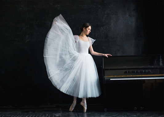 Ballerina im weißen Tutu stützt sich auf ein Klavier im dunklen Bühnenlicht