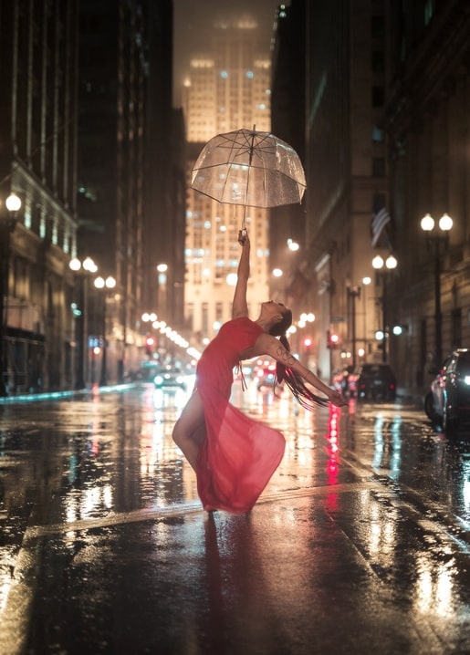 Ballerina im roten Kleid balanciert bei Regen nachts unter einem durchsichtigen Regenschirm
