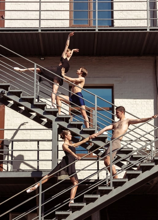 Vier Balletttänzer springen auf einer Außentreppe vor weißer Backsteinfassade