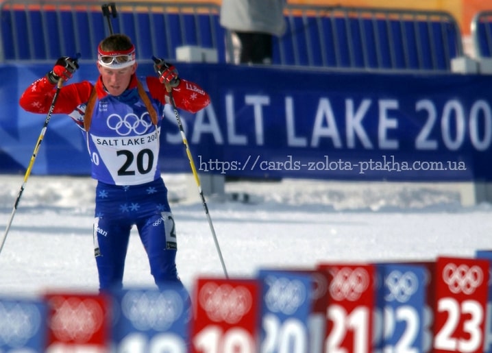Біатлоніст з піднятими лижними палицями мчить на тлі олімпійських кілець