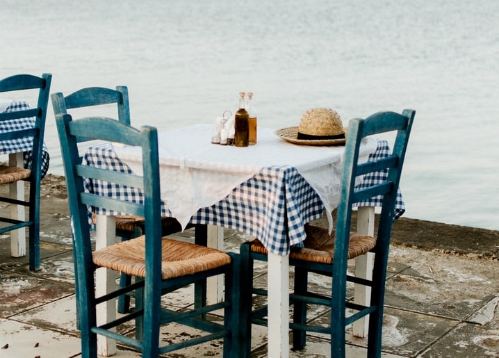 Blue wooden chairs around a table with a checkered cloth and straw hat by the sea