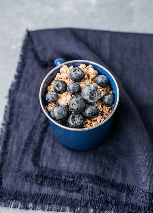 Heidelbeeren auf Granola in einer blauen Emaille-Tasse auf dunkelblauem Leinen