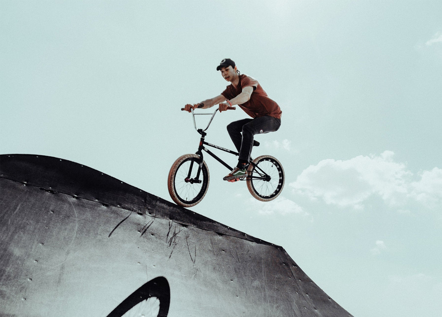 BMX-Fahrer im orangefarbenen Shirt springt mit dem Rad von einer Stahlrampe vor bewölktem Himmel