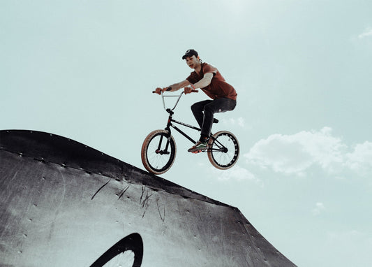 BMX-Fahrer im orangefarbenen Shirt springt mit dem Rad von einer Stahlrampe vor bewölktem Himmel