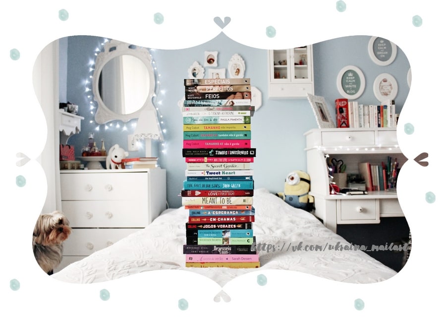 Tall stack of books on a bed with a small Yorkshire terrier sitting on the floor of a light blue bedroom