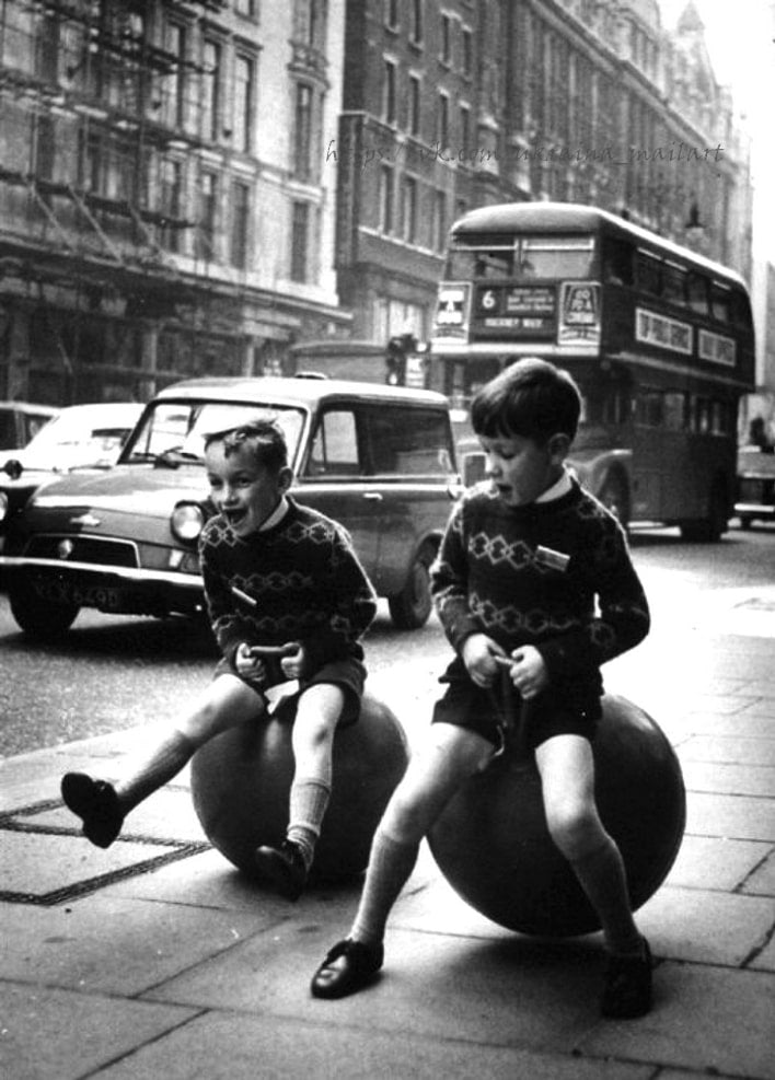 Vintage photo of two boys bouncing on space hoppers on a London sidewalk with a red double-decker bus behind them