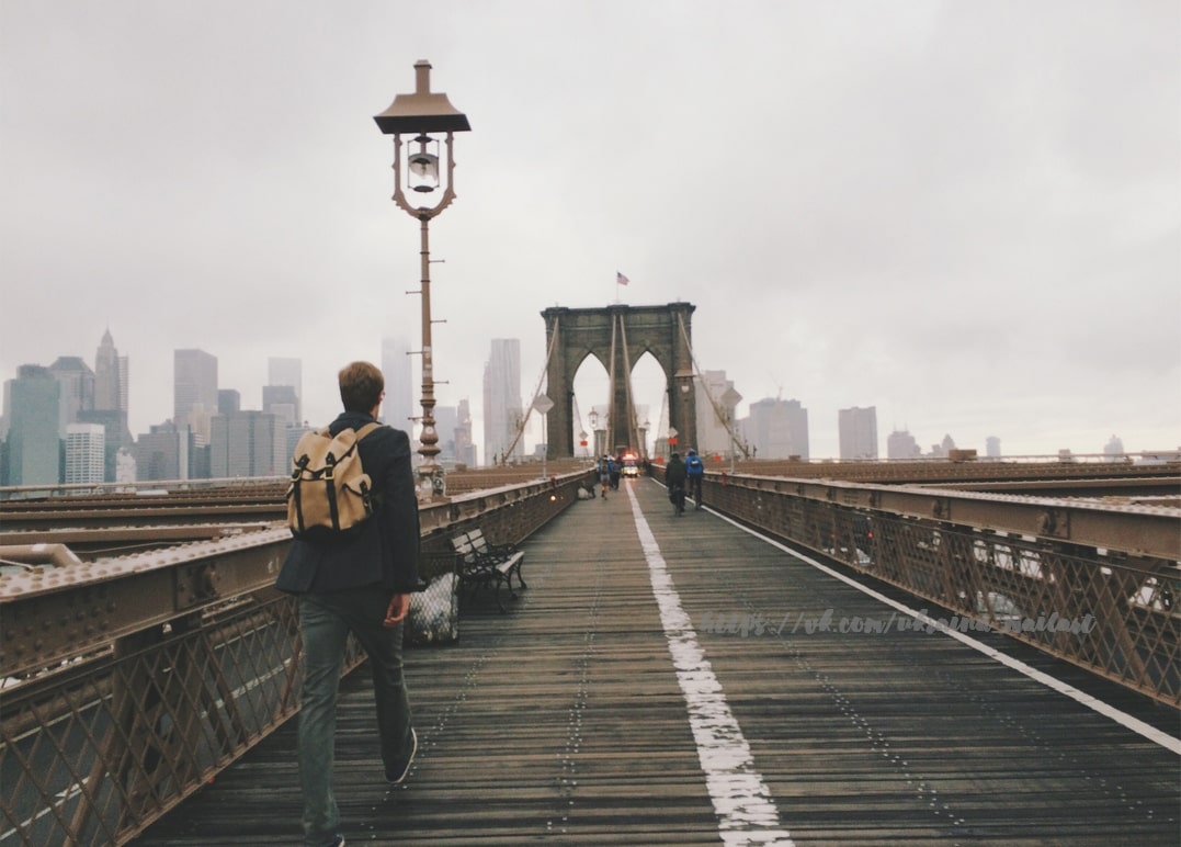 Man with a backpack walking across the Brooklyn Bridge toward the New York skyline
