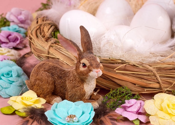 Brown bunny figurine beside a nest of white eggs with pastel fabric flowers and feathers